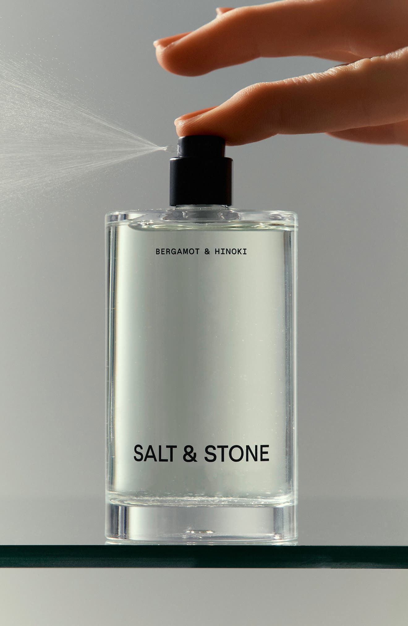 Bottle of Salt & Stone fragrance being sprayed with a hand against a neutral background