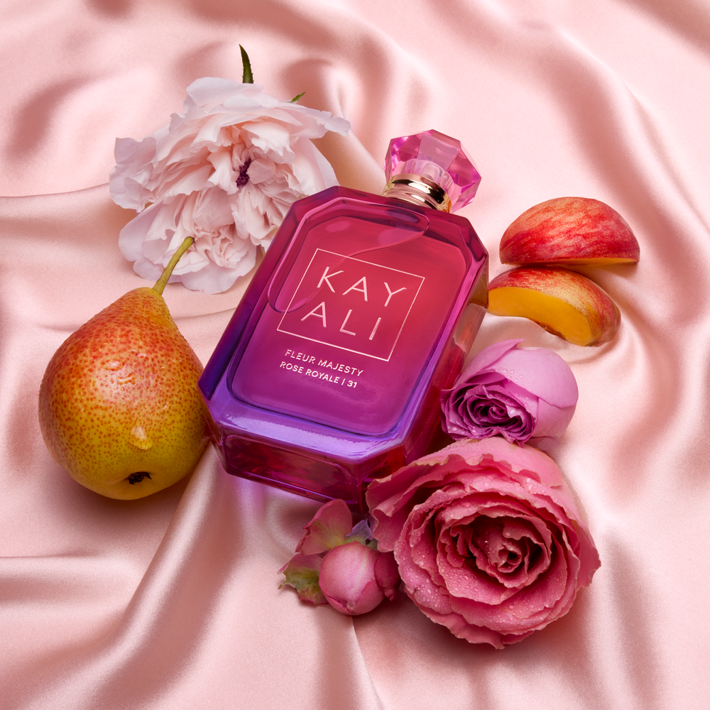 KAYALI perfume bottle surrounded by flowers and fruits on a pink fabric background